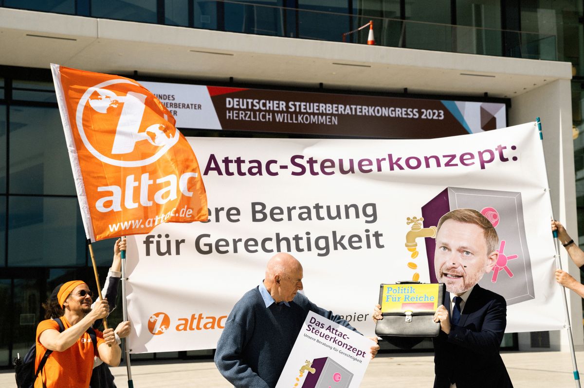 Vor dem Deutschen Steuerberaterkongress stehen Aktivist*innen mit einer Attac-Fahne und einem Transparent mit dem Slogan "Das Attac-Steuerkonzept: Unsere Beratung für Gerechtigkeit", davor kabbeln sich ein älterer Attac-Aktivist und ein Mensch mit Christian-Lindner-Maske