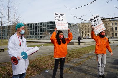 Ein Aktivist ist als Arzt verkleidet und hält eine übergroße Spritze, auf der Solidarität steht. Daneben halten Aktivist:innen Schilder.