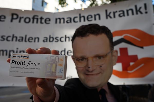 Mensch mit Jens Spahn-Maske posiert mit einer Fake-Medikamentenpackung "Profit forte"