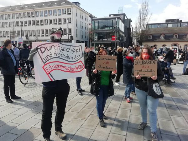 Eine Gruppe Attacis mit Schildern, einer ist als Echsenmensch verkleidet und hält ein Schild mit der Aufschrift "Durchgechippt und trotzdem da: Echsenmenschen-Antifa"