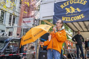Auf dem Lautsprecherwagen steht ein Man mit Mikrophon und oranger Jacke und hält einen orangen Regenschirm auf dem "Schutzschirm" steht, hinter ihm hängt eine Refugees-welcome-Flagge