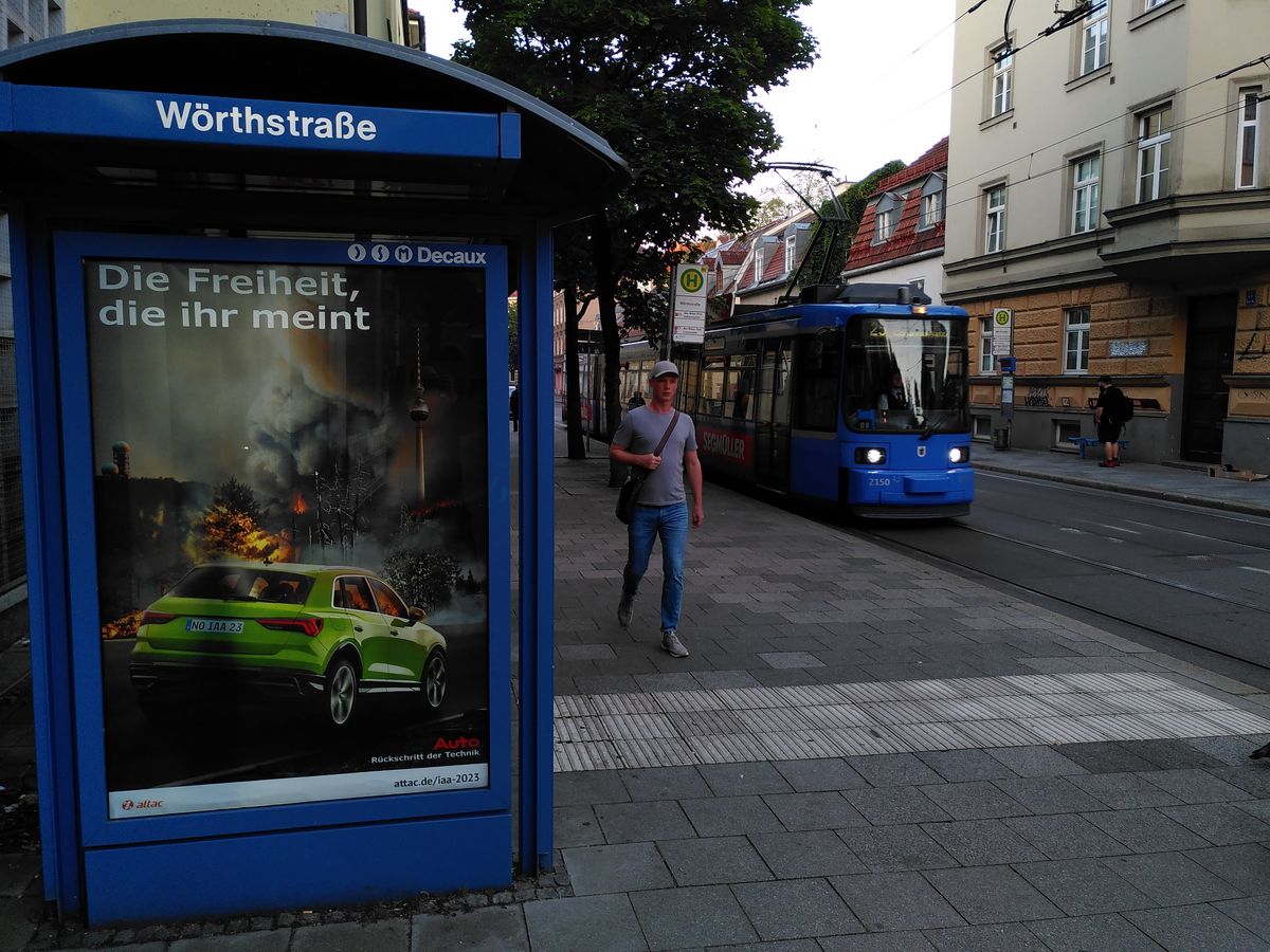 in einer Straßenbahnhaltestelle in München hängt ein gefasstes Werbeplakat, dass einen großen SUV vor einem im Waldbrand versinkenden Berlin zeigt und der Überschrift "Die Freiheit, die ihr meint", daneben fährt eine Straßenbahn ein