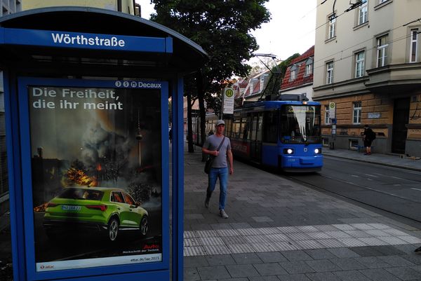 Links ist eine Straßenbahnhaltestelle mit einem Adbustingplakat zu sehen, das einen SUV vor einem brennenden Berlin zeigt und dem Slogan "Die Freiheit, die ihr meint", rechts fährt eine Straßenbahn