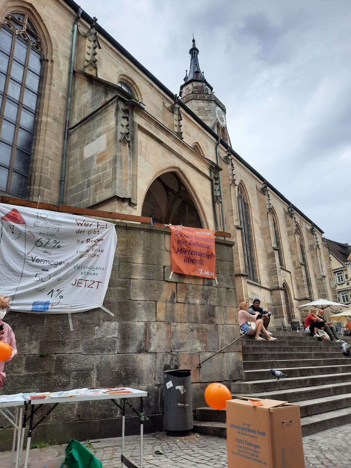 Im Bild ist ein Infostand von Attac in Tübingen, im Vordergrund ist ein Umzugskarton, der für das Vermögen der ärmeren Bevölkerungshälfte steht und im Hintergrund der Kirchturm der Marktkirche, die 67m Hoch ist und damit das Vermögen der reichsten 10% als Vergleich darstellt.