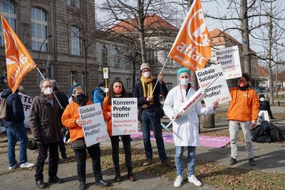 Ein Aktivist ist als Arzt verkleidet und hält eine übergroße Spritze, auf der Solidarität steht. Im Hintergrund halten Aktivist:innen Schilder.