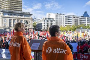 Zwei Frauen mit orangen Attac-Jacken sind von hinten auf einer Bühne zu sehen, dahinter ist ein Blick über den vollen Kundgebungsplatz.