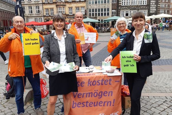Bild von einem Infostand von Attac auf dem Marktplatz in Bremen, zu sehen sind Aktivist*innen in orangen Jacken und davor zwei Frauen im Buiseneskostüm mit 100€ Scheinen, dazu ein kleines Plakat auf dem "Ich habe, also gebe ich" steht.