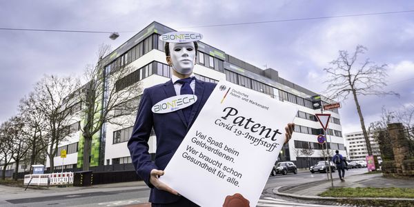 Vor der Biontech-Firmenzentrale steht ein Mensch im Anzug mit Maske und Biontech-Logo am Revers, der eine überdimensionierte Patenturkunde hält