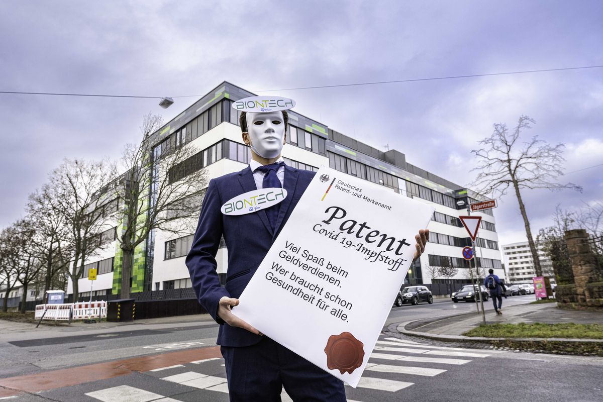 Vor der Biontech-Zentrale steht ein Mensch im Anzug mit Maske und dem Biontech-Logo am Revers und einer überdimensionierten Patenturkunde in der Hand.