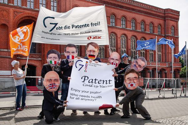 Vor dem Roten Rathaus in Berlin stehen als G7-Gesundheitsminister verkleidete Aktivist*innen, die eine übergroße Patenturkunde auf Impfstoffe in den Händen halten. Hinter ihnen fordern Attac-Aktivist*innen auf einem Transparent auf die Patente freizugeben. Im Hintergrund sieht man G7-Flaggen 