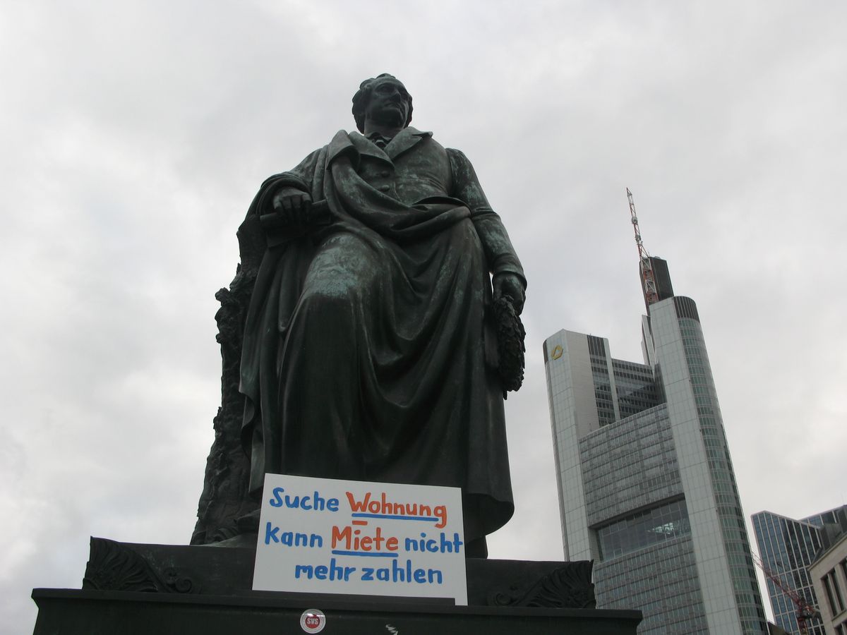 Bild vom Goethedenkmal in Frankfurt, am Fuß der Statue steht ein Schild mit der Aufschrift "Suche Wohnung, kann Miete nicht mehr zahlen"