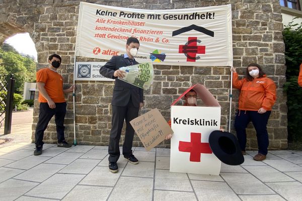 Ein Man im Anzug mit Logos privater Klinikkonzerne und Geldscheinen steht neben einem Menschen im Kostüm eine Kreisklinik, die Bettelt, im Hintergrund das Aktionstransparent.