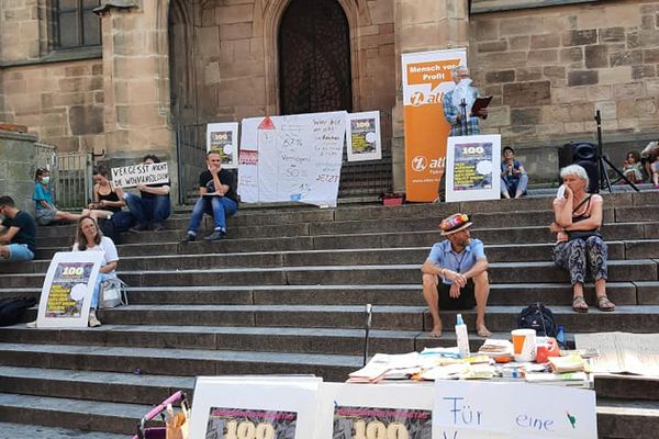 Auf der Treppe vor einer Kirche sitzen Menschen für eine Kundgebung, im Vordergrund ist ein Infostand mit einem Plakat auf dem "Für eine Vermögenssteuer! Aber was ist mit unserem Häusle? Freibetrag 1 Mio. Euro" steht