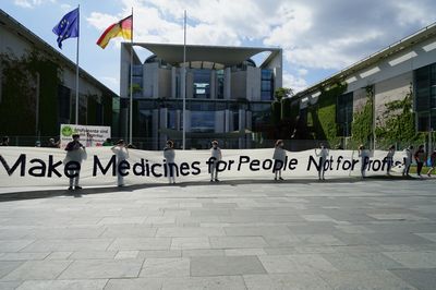 Vor dem Bundeskanzlerinnenamt halten mehrere Menschen ein ca. 15m Langes Transparent mit dem Slogan "Make Medicines for People Not for Profit"