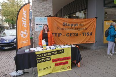 Ein Infostand mit einer Attac-Beachflag und einer Aktivistin, hinter ihr ist ein Transparent mit dem Slogan "Stoppt die Freihandelsagenda: TTIP – CETA — TISA"