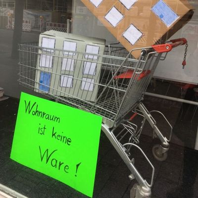 Ein Schaufenster, in dem ein Einkaufswagen, der mit Häusern gefüllt ist steht, davor ist ein Schild mit der Aufschrift "Wohnen ist keine Ware!"