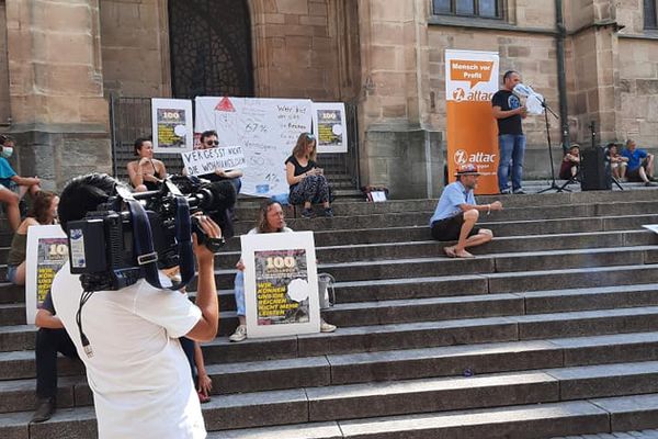 Auf den Stufen vor einer Kirche sitzen Menschen mit Transparenten und Plakaten von Wer hat der gibt, im Vordergrund ist ein Kameramann zu sehen