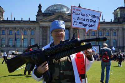 Vor dem Reichstag steht eine Aktivistin in einer Camouflage-Uniform und darüber ein Pflegekittel und eine OP-Haube, in ihren Händen hält sie ein aufblasbares Maschinengewehr und ein Schild mit der Aufschrift "Muss ich schießen, damit es Geld für Pflege gibt?"