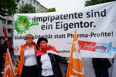 Zwei Aktivistinnen stehen vor einem Transparent mit der Aufschrift "Impfpatente sind ein Eigentor. Globale Solidarität statt Pharma-Profite!"