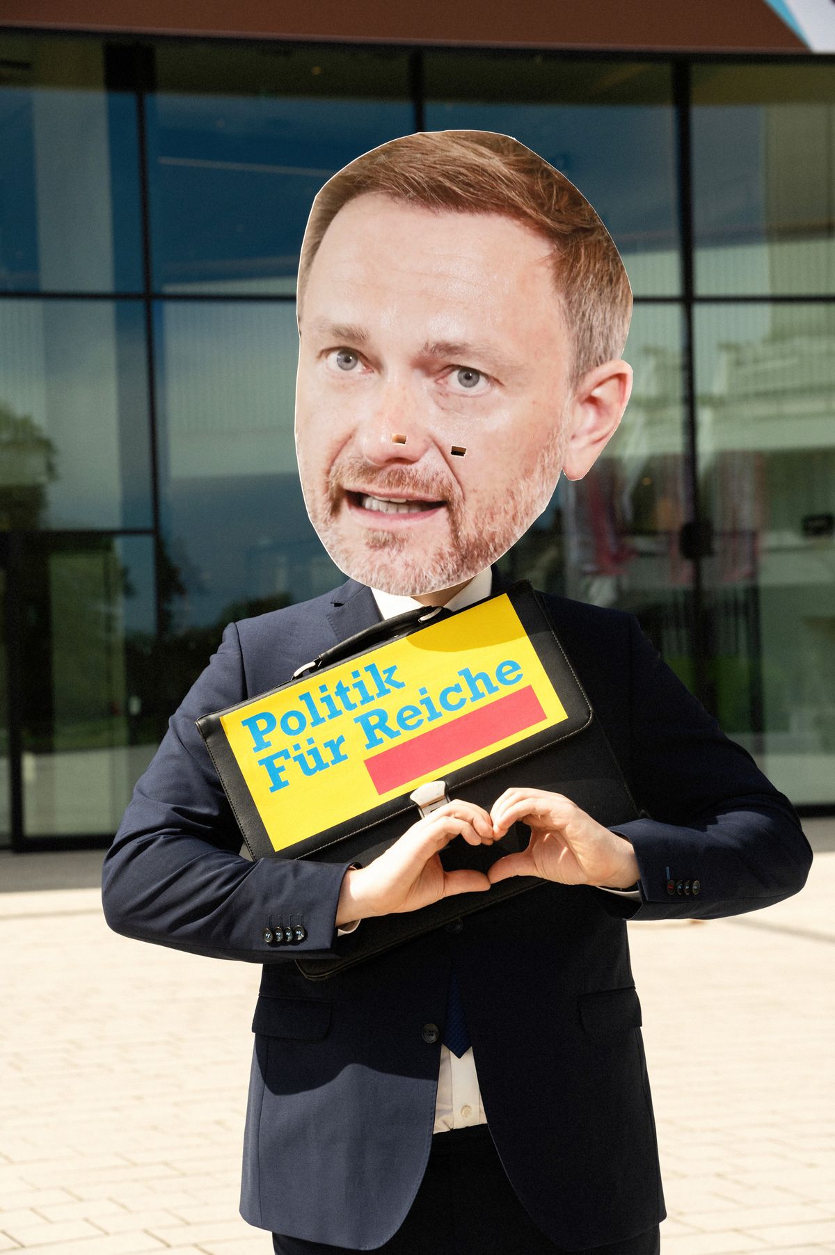 Ein Mensch mit Christian-Lindner-Maske hält eine Aktentasche, auf der im Stil des FDP-Logos "Politik für Reiche" steht, beide Hände sind so zusammengelegt, dass sie ein stilisiertes Herz bilden