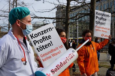 Ein Aktivist ist als Arzt verkleidet und hält eine übergroße Spritze, auf der Solidarität steht. Im Hintergrund halten Aktivist:innen Schilder.