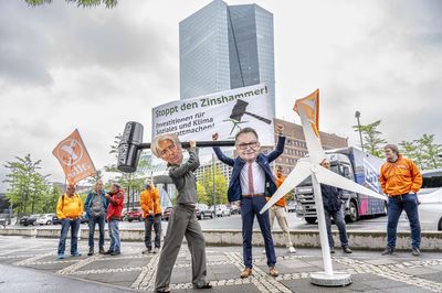 Die als Lagarde verkleidete Aktivistin holt aus um mit dem Zinshammer auf ein Windrad einzuschlagen, im Hintergrund stehen die Attac-Aktivist*innen vor dem EZB-Turm