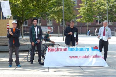 Im Bild ist ein Infostand mit Attacies, im Vordergrund ist ein Plakat, das die Vermögensverteilung in Deutschland mit der Höher der dahinter liegenden Reinholdikirche Vergleicht und erklärt, dass die reichsten 10 Prozent 67 Prozent des Gesamtvermögens besitzen, während die ärmere Hälfte gerade ein Prozent besitzt.