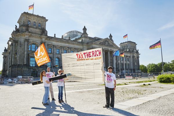 Zwei Aktivist*innen halten ein großes Banner auf der eine überdimensionierte Unterschriftenliste für Tax the Rich aufgemalt ist, eine Person unterschreibt mit einem überdimensionierten Füller. Im Hintergrund ist das Reichstagsgebäude zu sehen.