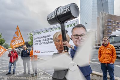 Aktivist*innen von Attac stehen mit einem Transparent mit dem Titel "Stoppt den Zinshammer! Investitionen für Klima und Soziales nicht plattmachen" vor der EZB. Vor ihnen haben sich zwei als EZB-Präsidentin Lagarde und Bundesbankchef Nagel verkleidet, die einen großen Hammer mit der Aufschrift Zinshammer tragen.