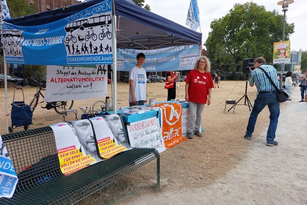 Bild von einem Infostand, neben dem eine Frau im roten T-Shirt steht und vor dem Infostand hängen Plakate mit einem Photo von Christian Lindner und dem Slogan "Kein Geld für ÖPNV? Sollen sie doch Porsche fahren."