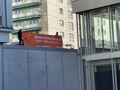 Auf dem Dach eines Nebengebäudes stehen drei Aktivisten die ein Banner mit dem Slogan "Power to the people – Fossillobby entmachten!" steht