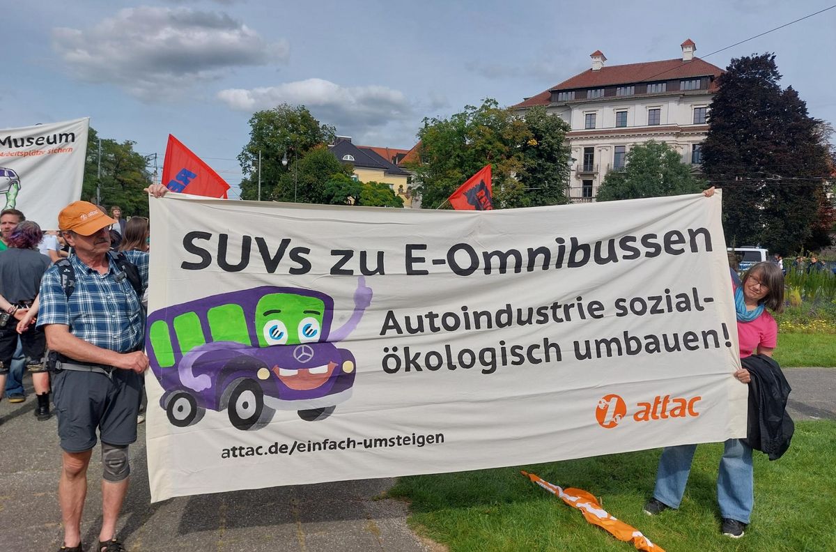 zwei Attacies halten ein Transparent. Darauf die Forderung "SUVs zu E-Omnibussen - Autoindustrie sozialökologisch umbauen" und Attac-Logo mit Webseitenangabe attac.de/einfach-umsteigen