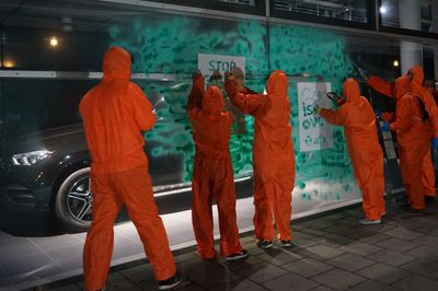 Menschen in orangen Overalls stehen vor einem grün angesprühtem Autohausschaufenster und legen Schablonen an um Text darauf zu sprühen.