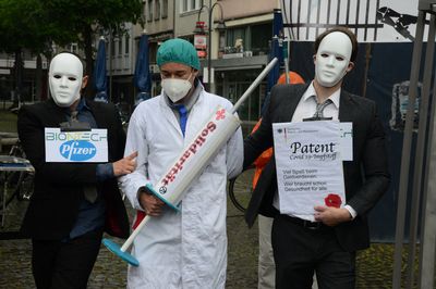 Ein als Arzt verkleideter Mensch mit einer Spritze, auf der Solidarität steht, wird von zwei Menschen im Anzug, die die Pharmaindustrie symbolisieren abgeführt.