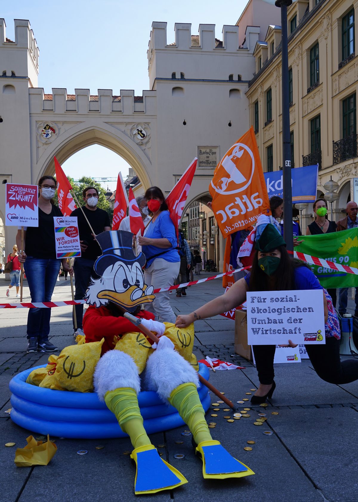 Eine als Dagobert Duck verkleidete Person sitzt in einem Swimmingpool voll Geldsäcken, während eine als Robin Hood verkleidete Person einen Geldsack entreißt. Im Hintergrund sind Demonstrant:innen zu sehen.
