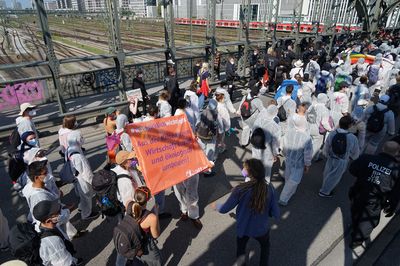 In einem Demoblock mit Menschen mit weißen Maleranzügen, der über eine Brücke im Gleisvorfeld des Münchener Hauptbahnhofes läuft, hält ein Mensch eine Fahne mit dem Text "Aus der Krise lernen: Wirtschaft sozialökologisch umbauen!"