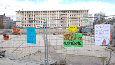 An einem Bauzaun vor einer Brache sind mehrere Protestschilder angebracht.