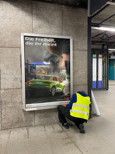 Ein Aktivist in gelber Warnweste hängt in einem U-Bahnhof ein gefasstes Werbeplakat auf, auf dem ein dicker SUV vor einem in Waldbränden erstickten Berlin fährt und dem Slogan "Die Freiheit, die ihr meint."