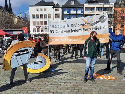 Attacis halten ein Banner mit dem Slogan "Vonovia-Dividende: 38 Cent von jedem Euro Miete" davor reißt ein als Vermieter Verkleideter Aktivist ein gutes Drittel aus einer überdimensionierten Euromünze, der die Miete symbolisiert