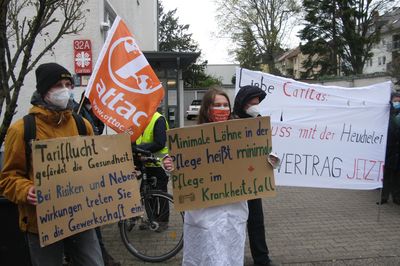 Zwei Attacis halten Schilderfür bessere Pflegebedingungen in die Kamera, im Hintergrund ist die Diakonie.