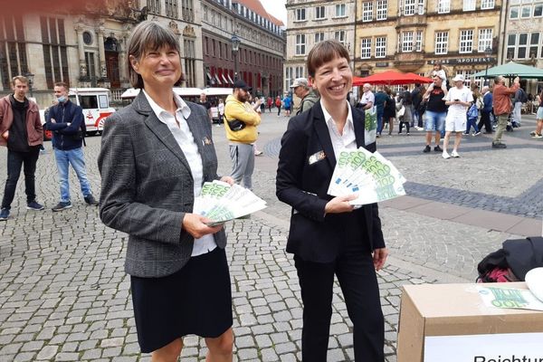 Zwei Frauen im Buiseneskostüm stehen auf dem Bremer Marktplatz und halten Flyer, die wie 100€ Scheine aussehen gefächert in ihrer Hand. 
