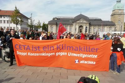 Auf einem Platz in einer Kundgebung halten Menschen ein großes oranges Transparent mit der Aufschrift "CETA ist ein Klimakiller. Stoppt Konzernklagen gegen Umweltgesetze"