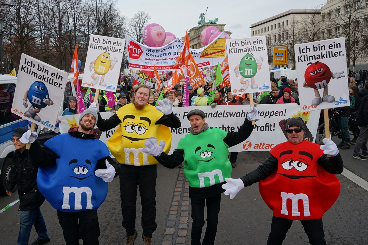 Vier als M&M verkleidete Aktivist*innen halten Schilder auf denen ein M&M abgebildet ist und der Slogan "Ich bin ein Klimakiller" hinter ihnen ist der Attac-Demoblock mit einem Transparent gegen klimaschädliche Palmöle in Schokolade