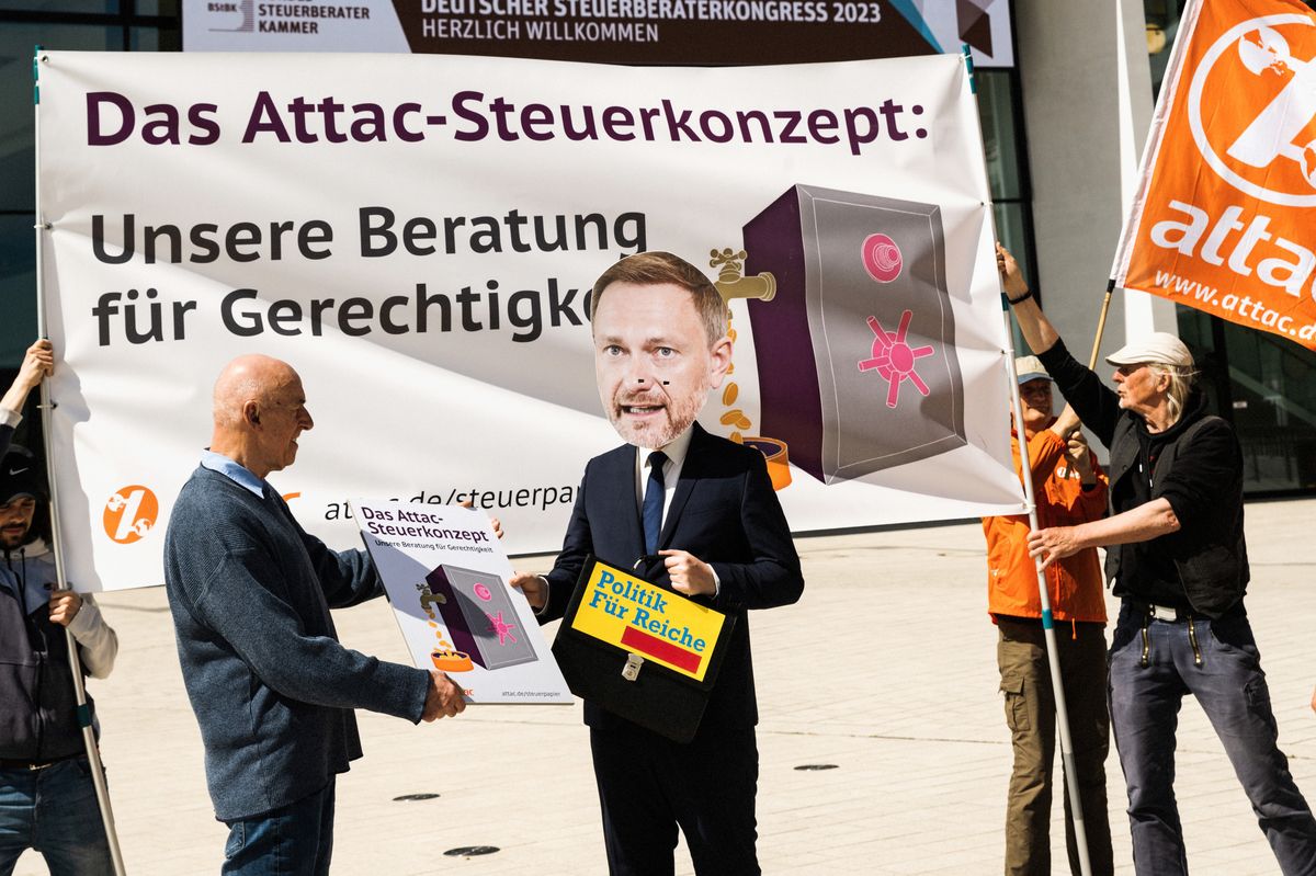 Ein Mensch mit Lindner-Maske und einer Aktentasche mit einem FDP-Logo, bei der der Parteiname durch den Slogan "Politik für Reiche" ersetzt wurde, bekommt das Attac-Steuerkonzept überreicht. Im Hintergrund halten zwei Aktivisten ein Banner mit der Aufschrift "Das Attac-Steuerkonzept: Unsere Beratung für Gerechtigkeit"