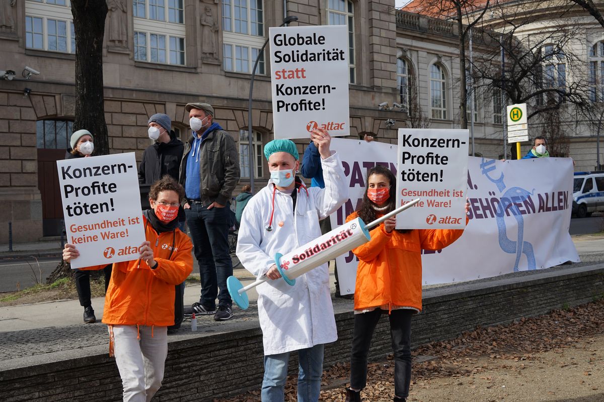 Ein Aktivist ist als Arzt verkleidet und hält eine Spritze mit der Aufschrift "Solidarität" im Hintergrund Aktivist:innen mit Schildern und Transparente zu sehen