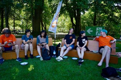 Attacies sitzen auf zu Bänken umgelegten Bierbänken und unterhalten sich, im Hintergrund sind Bäume zu sehen.