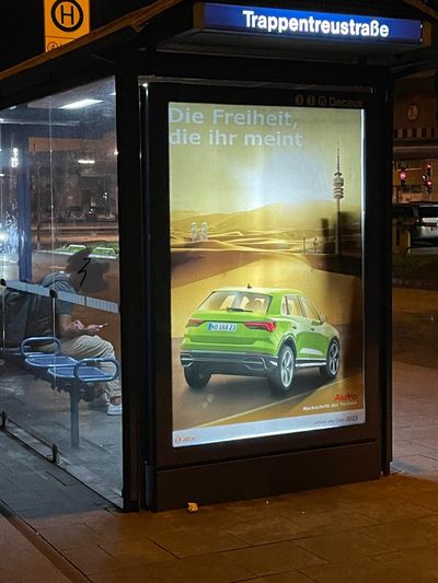 In einem Werbeschaukasten hängt ein gefaktes Plakat, das wie eine Autowerbung aussieht, in der ein dicker SUV vor einem in der Wüste versunkenen München fährt, mit dem Slogan "Die Freiheit, die ihr meint"