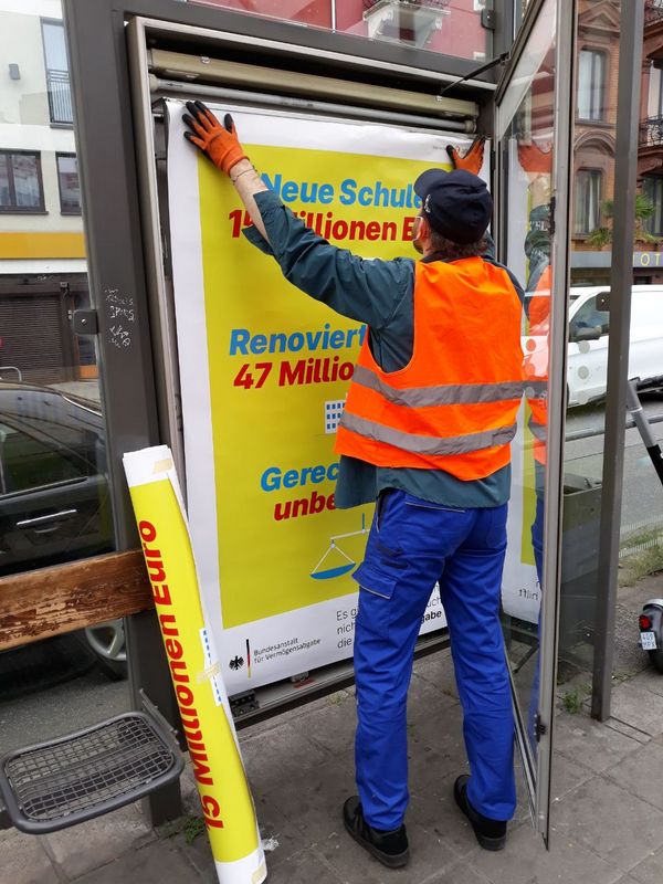 Eine Person in Warnweste hängt Plakate in einen Werbekasten