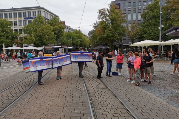 Drei Menschen stehen in einem tragbaren Straßenbahnkostüm, neben ihnen stehen Menschen zum Unterschreiben für das 9€-Ticket an.