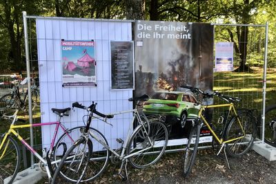 Vor einem Bauzaun stehen Fahrräder, am Bauzaun hängt ein City-Light-Poster auf dem eine Autowerbung persifliert wird